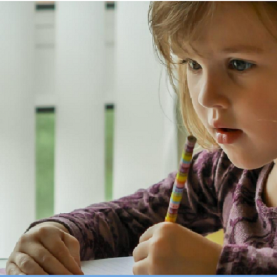 A young girl with blonde hair holding a rainbow coloured pencil in her right hand  drawing on white paper.