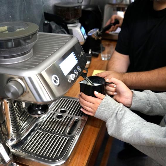 A young person holding a milk jug under a coffee machine to froth the milk