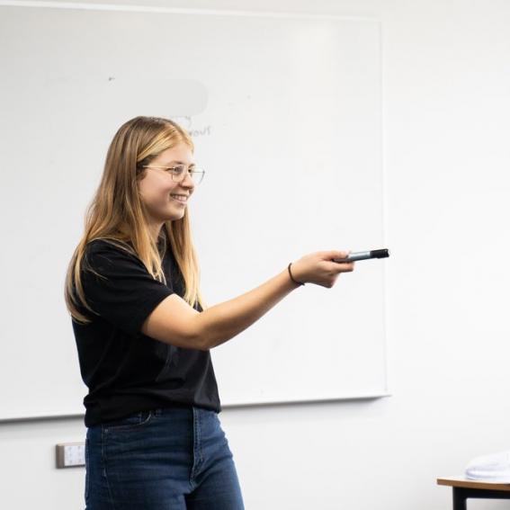 A young woman with long blond hair stands in front of a whiteboard holding out a whiteboard marker