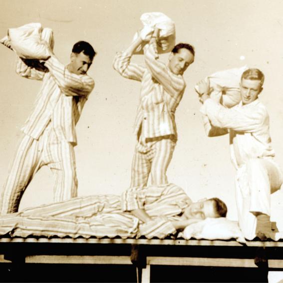 Old timey photo of four men on a roof throwing pillows at each other.