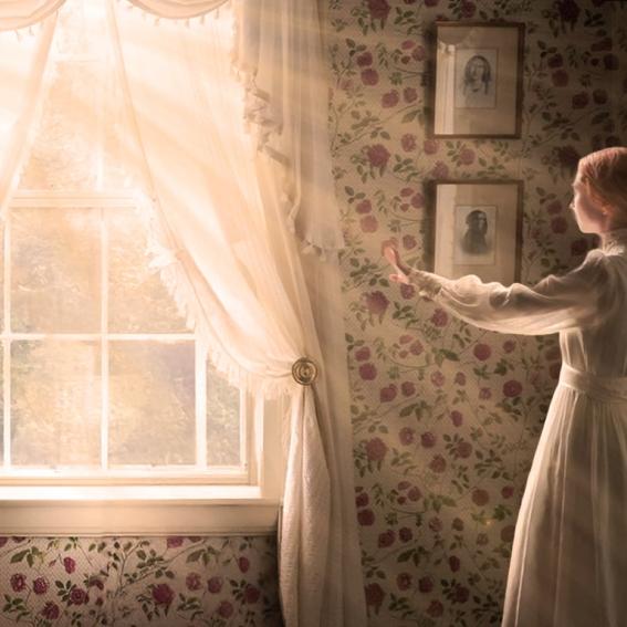 A young woman in Victorian dress stretches out a hand towards the light streaming in from the large window before her