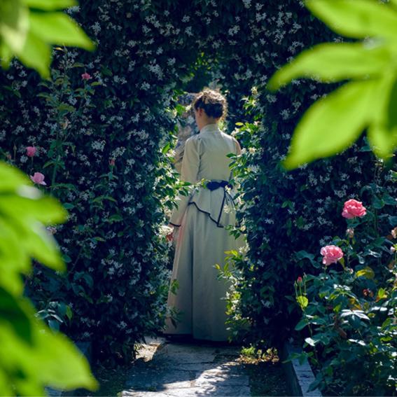 Woman dressed in an 18th century outfit walking through a rosebush garden.