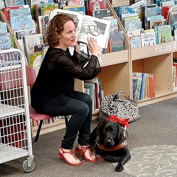 woman in black sitting on a chair in the library with her guide dog at her feet