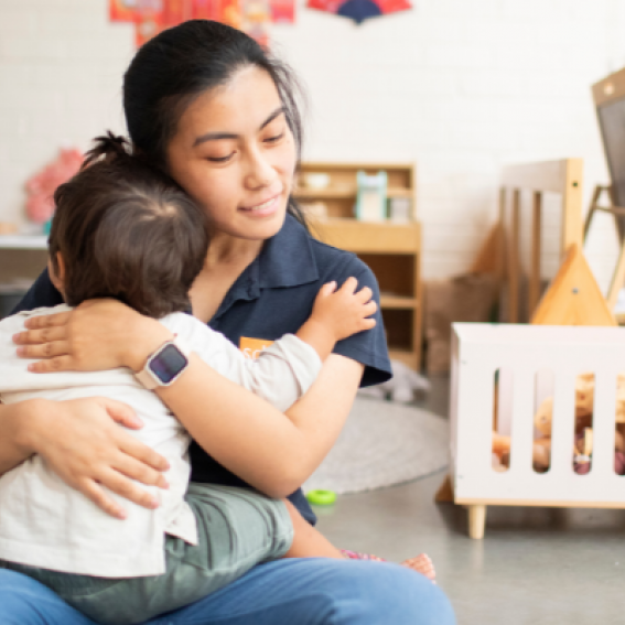 An educator at SDN Mosman warmly embracing an infant child in their indoor area.