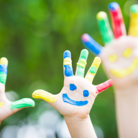 Two children's hands with their fingers painted yellow, green, blue and red with a blue smiley face painted on their palm.