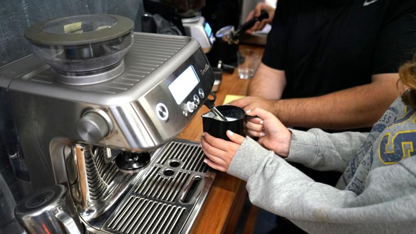 A young person holding a milk jug under a coffee machine to froth the milk