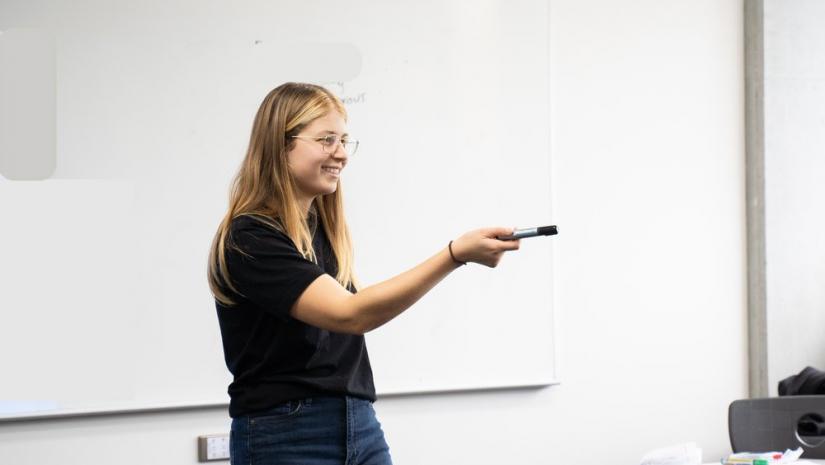 A young woman with long blond hair stands in front of a whiteboard holding our a whiteboard marker.