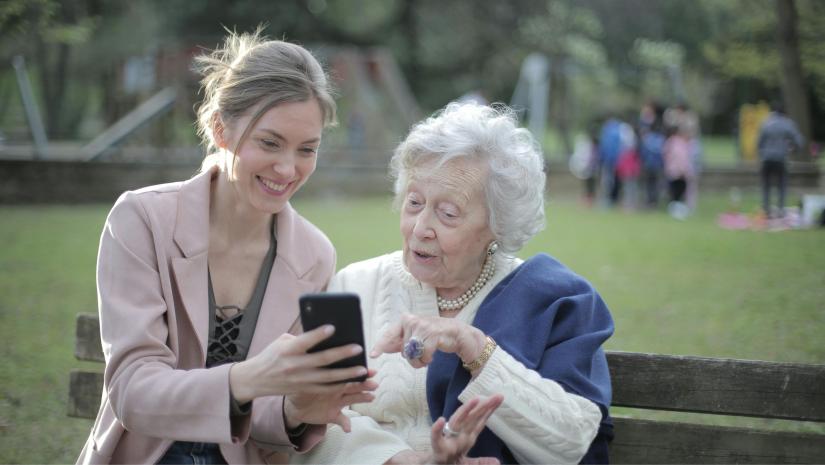 An elderly woman and younger woman sitting on a bench outside.