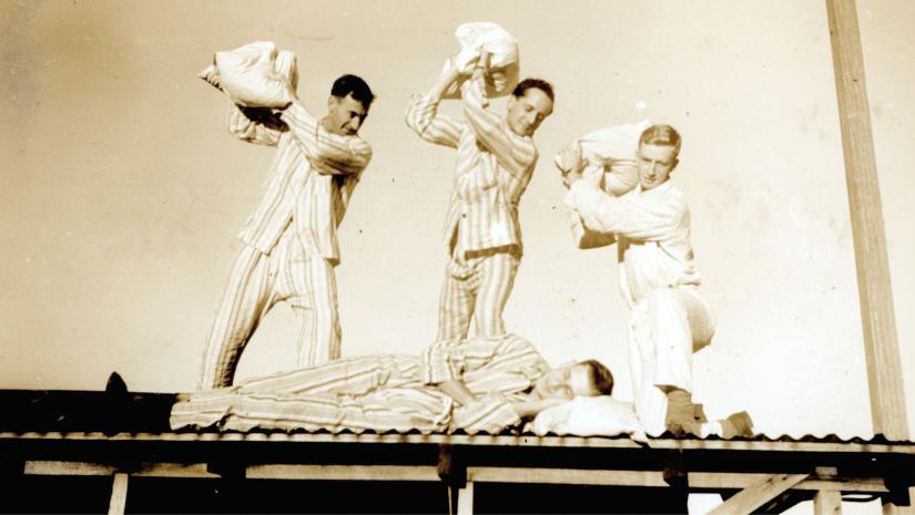 Old timey photo of four men on a roof throwing pillows at each other.