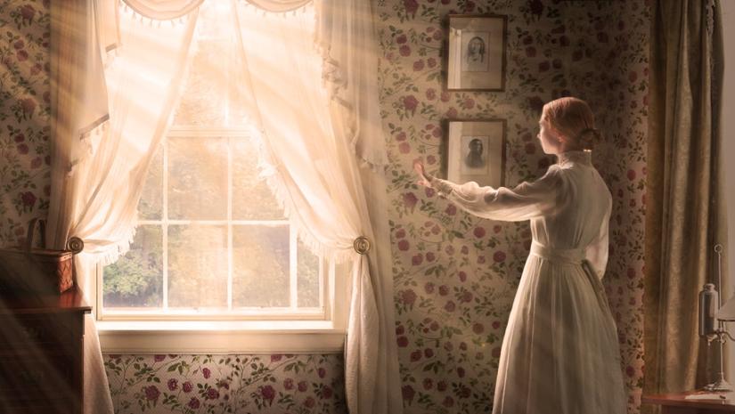 A young woman in Victorian dress stretches out a hand towards the light streaming in from the large window before her