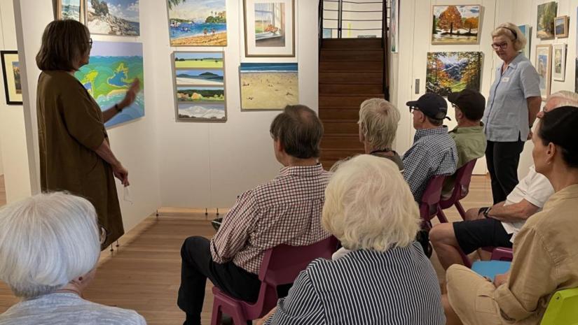 A group of people sitting infront of artworks in a gallery listening to a guide explain the works.