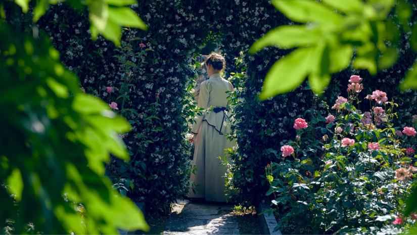 Woman dressed in an 18th century outfit walking through a rosebush garden.