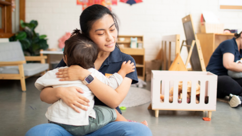 An educator at SDN Mosman warmly embracing an infant child in their indoor area.